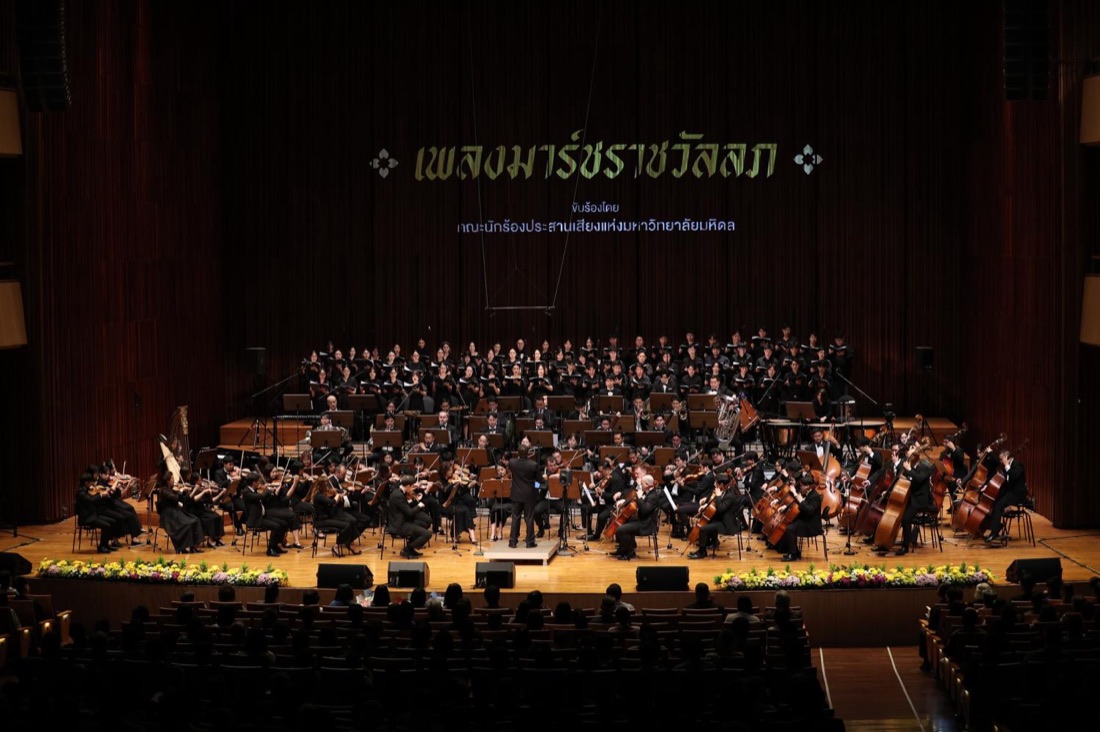 The Thailand Philharmonic Orchestra and chorus performing on the stage of Mahidol Siddhachada Hall, with the title of the Royal Guards' March projected in gold against the velvet backdrop.